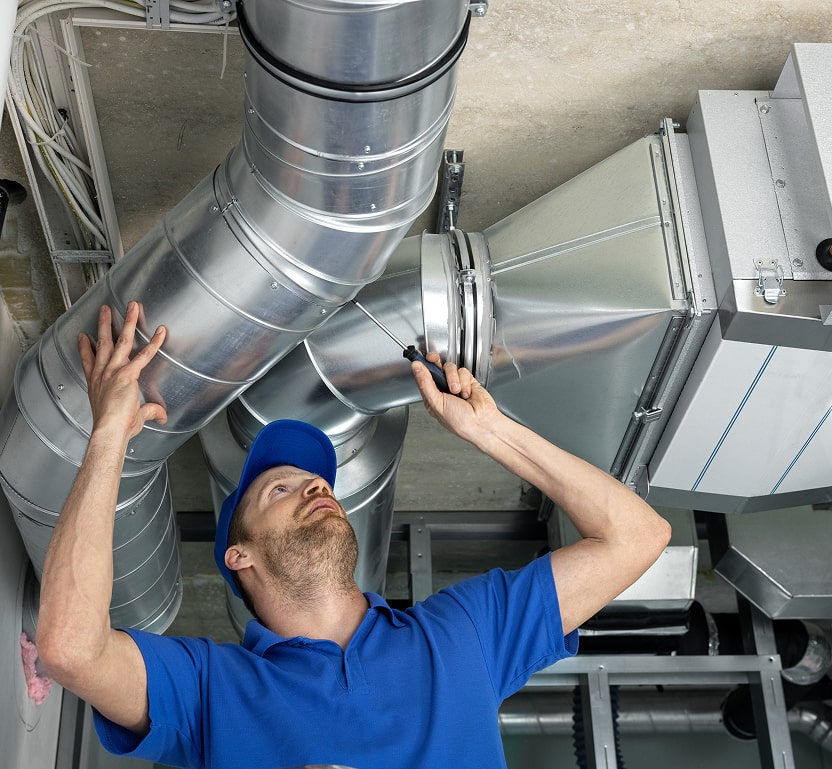 A worker inspecting ductwork near a ceiling, using a screwdriver on the area.