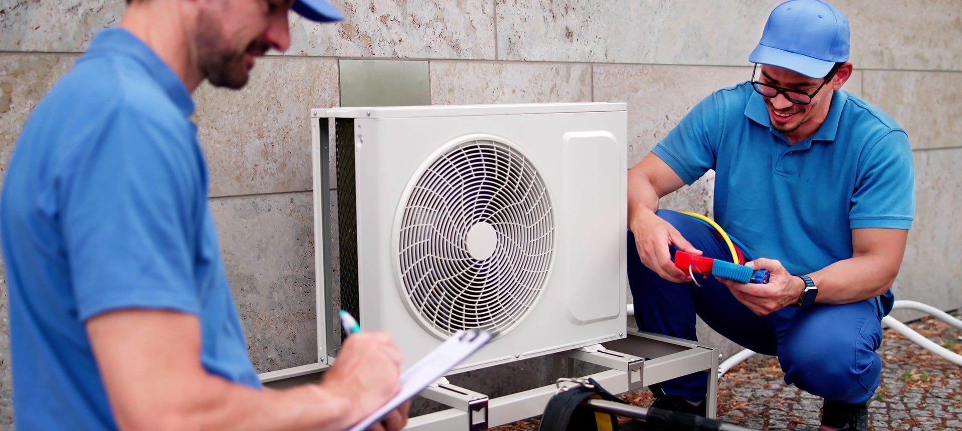 2 workers performing maintenance on an AC unit, one with tools, the other holding and writing on a clipboard.