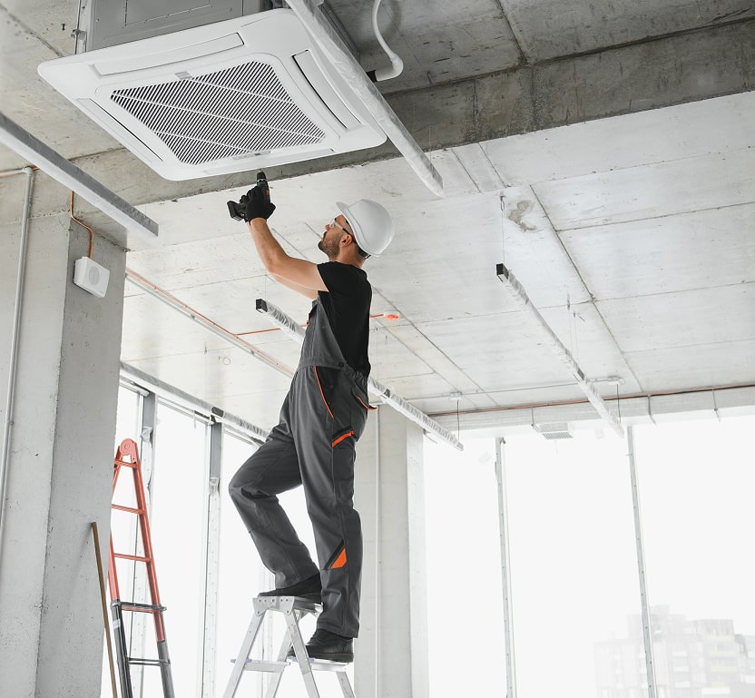 A worker standing on top of a ladder, using a drill on a vent system near a commercial building ceiling.